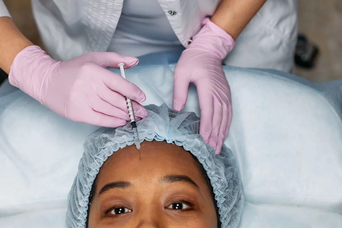 A gloved doctor injecting a patient's forehead, the patient wearing a hair cap