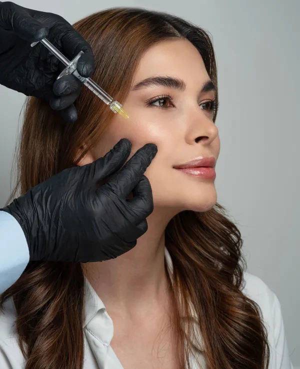 A woman receiving a filler injection in the cheek from a gloved doctor